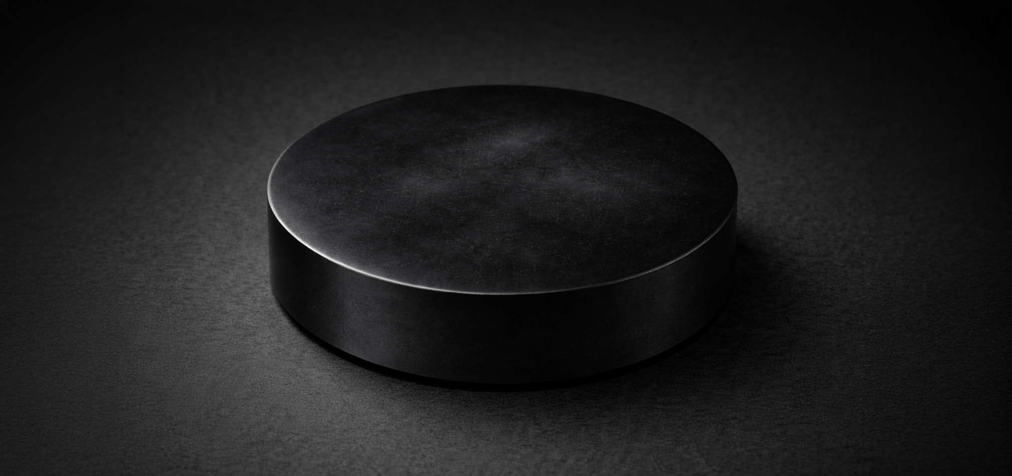 Studio detail view of the AGAR-7 microbial Host on a dark background, showing a sealed black petri-puck form with subtle off-centre bloom, particulate haze, and an optically withheld interior
